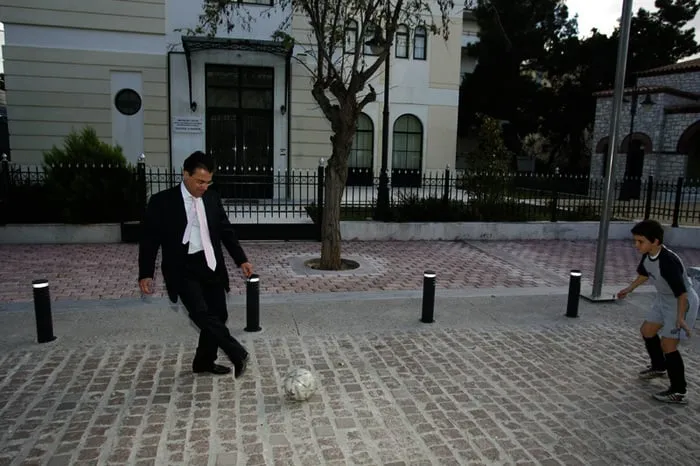 Ο Αργύρης Ντινόπουλος με κοστούμι κλωτσά μια μπάλα με ένα αγόρι σε πεζόδρομο.