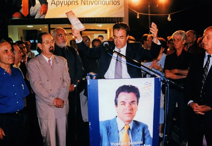 Νυχτερινή προεκλογική συγκέντρωση, ο Αργύρης Ντινόπουλος στο βήμα με υψωμένο χέρι.