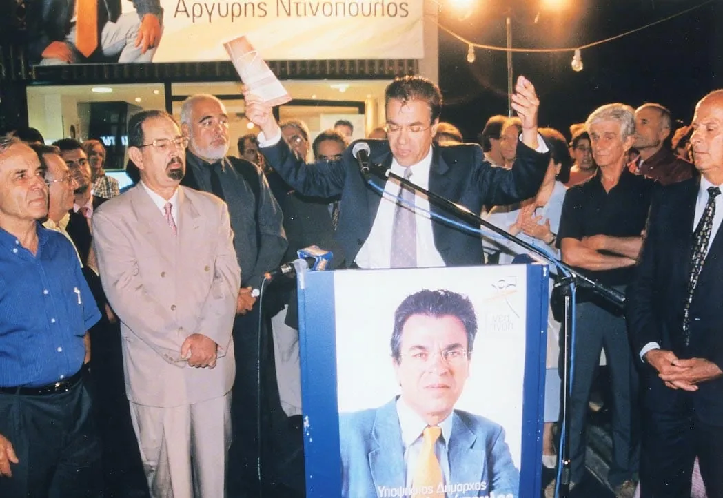 Νυχτερινή προεκλογική συγκέντρωση, ο Αργύρης Ντινόπουλος στο βήμα με υψωμένο χέρι.