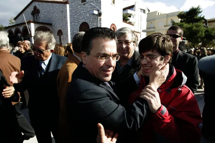 Ο Αργύρης Ντινόπουλος αγκαλιάζει έναν χαμογελαστό νεαρό άνδρα έξω από εκκλησία, με πλήθος γύρω τους.
