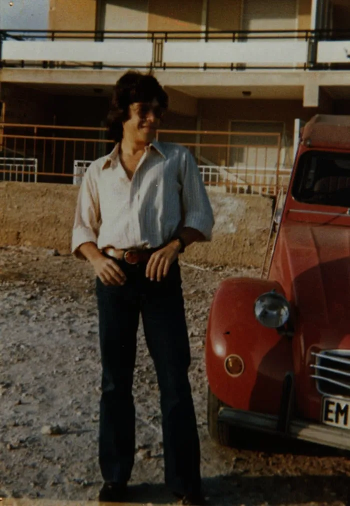 Νεαρός άνδρας στέκεται δίπλα σε ένα κόκκινο Citroën 2CV έξω από πολυκατοικία, δεκαετία 1970.