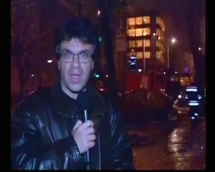 Ο Αργύρης Ντινόπουλος μιλά στην κάμερα νύχτα, με φωτισμένο κτίριο και οχήματα έκτακτης ανάγκης πίσω του, Βελιγράδι, 1999.