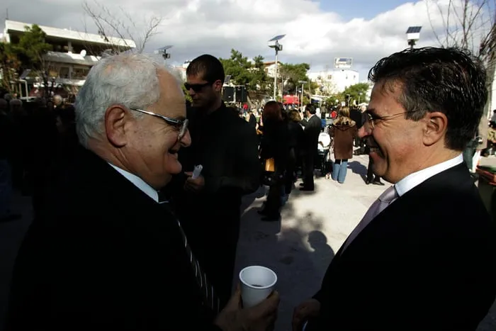 Ο Αργύρης Ντινόπουλος συνομιλεί χαμογελαστός με έναν άνδρα, ενώ ένας ηλικιωμένος κρατά καφέ σε γιορτινή πλατεία.