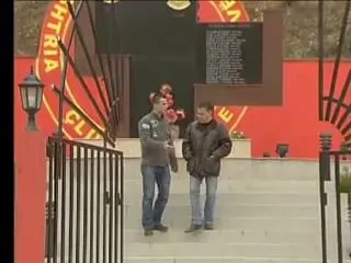 Κόκκινο μνημείο με μαύρη πλάκα που φέρει κατάλογο ονομάτων και κυκλικό έμβλημα· δύο άνδρες στα σκαλοπάτια, Κόσοβο, 2006.