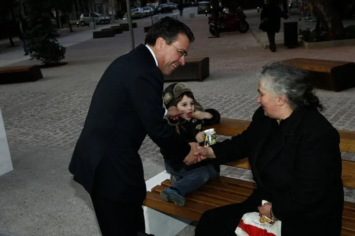 Ο Αργύρης Ντινόπουλος σκύβει να χαιρετήσει μια ηλικιωμένη γυναίκα σε παγκάκι, με ένα παιδί δίπλα της.