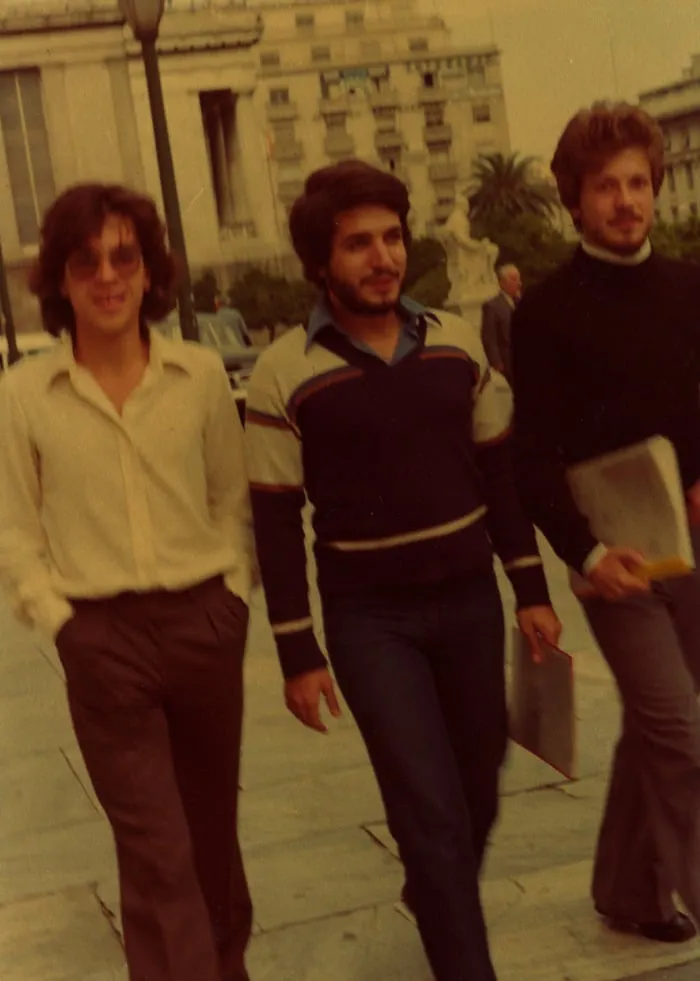 Σέπια φωτογραφία τριών νεαρών ανδρών που περπατούν κοντά σε ένα επιβλητικό κτίριο και ένα άγαλμα, μόδα της δεκαετίας του 1970.