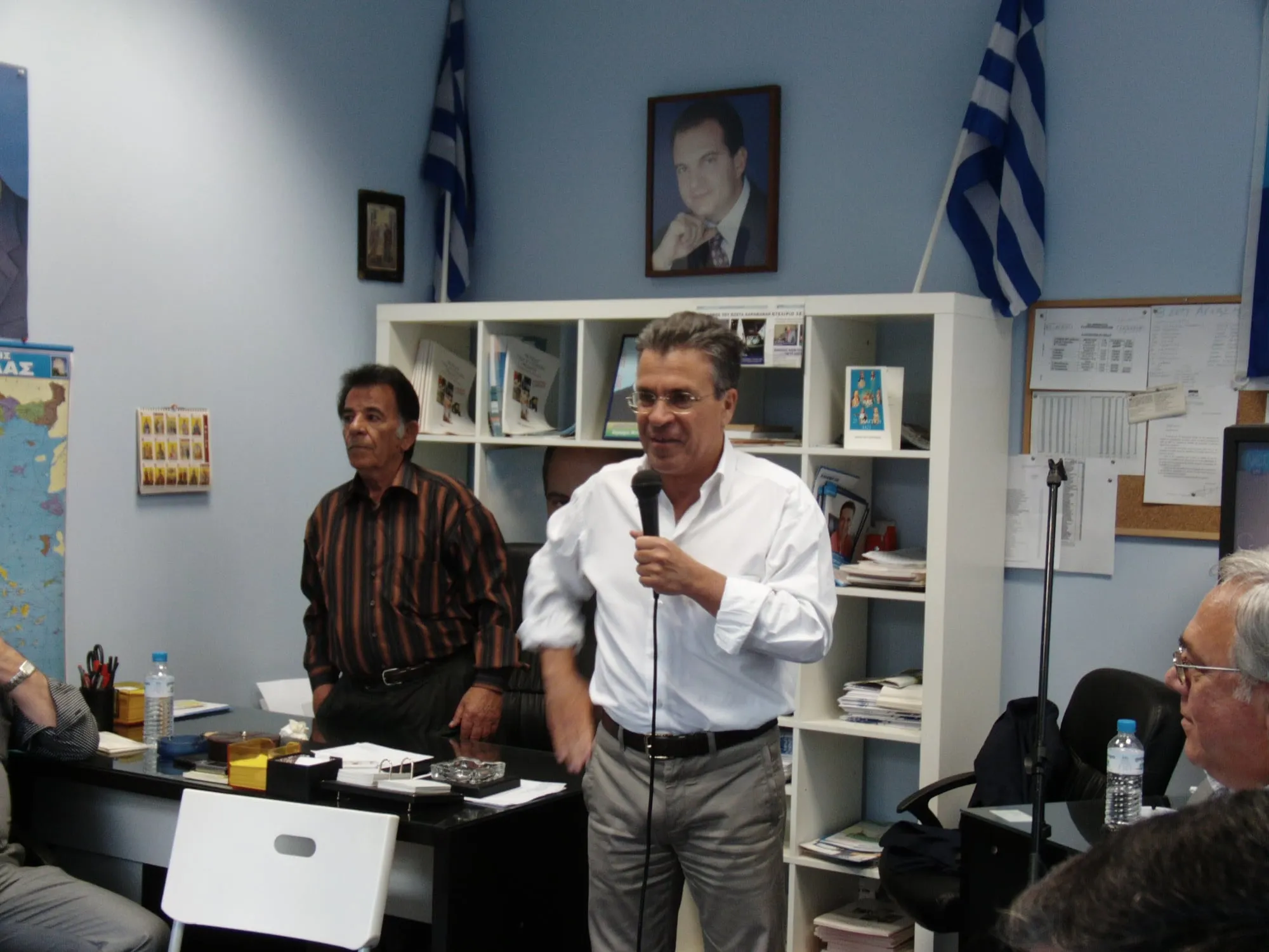 Ο Αργύρης Ντινόπουλος, με λευκό πουκάμισο και ανασηκωμένα μανίκια, μιλά κρατώντας μικρόφωνο σε γραφείο με γαλάζιους τοίχους, δύο ελληνικές σημαίες, κορνιζαρισμένο πορτρέτο άνδρα και λευκή ραφιέρα γεμάτη έντυπα· δίπλα του στέκεται ένας άνδρας και μπροστά κάθεται ακροατήριο.