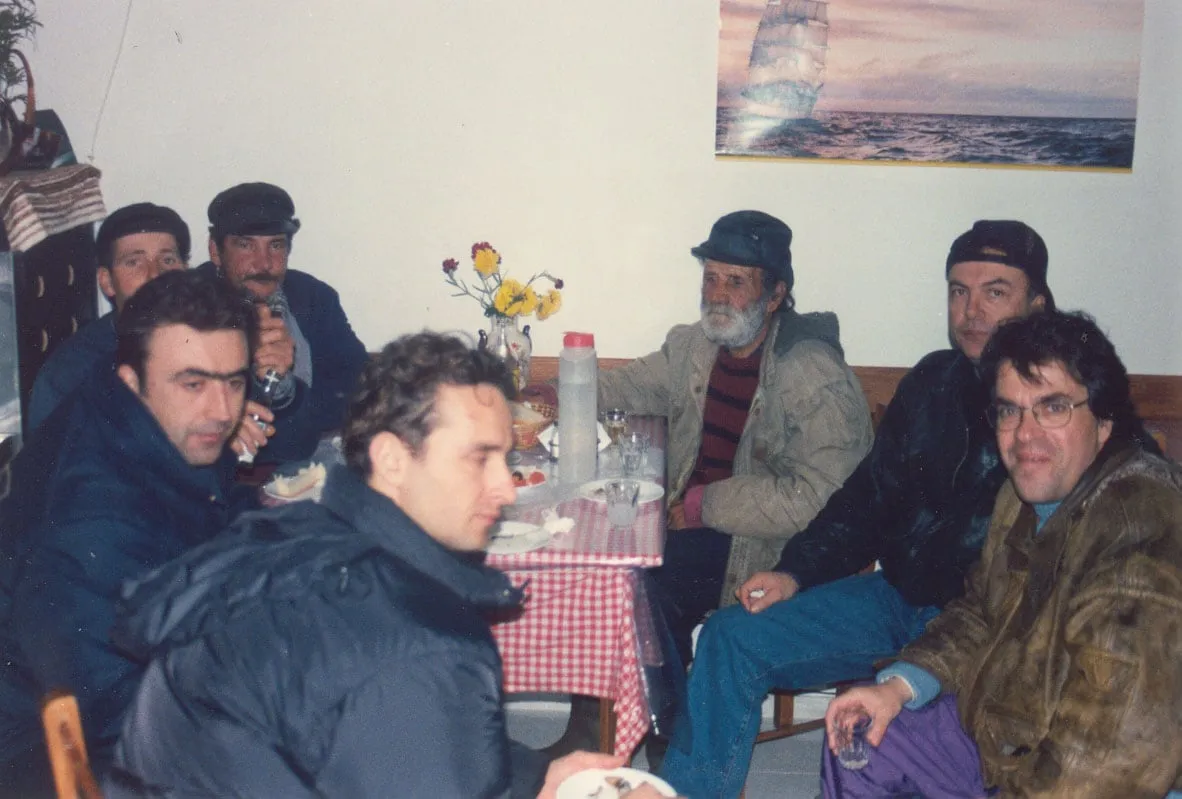 An interior with a pale wall. A group of men sit around a small table under a red checked cloth; on it a tall bottle, glasses, plates and a vase of red and yellow flowers. Several wear caps and heavy jackets; centre right, an older man with a white beard and a dark cap. On the wall behind them, a poster of a sailing ship on open sea. Bottom right, Argyris Dinopoulos, in glasses and a wide-collared jacket, holds a small glass and looks at the camera.