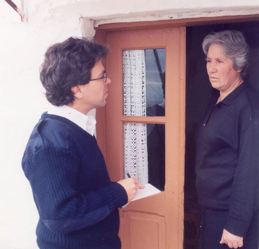 Two people at the doorway of a house with a whitewashed wall. At left, in profile, Argyris Dinopoulos in glasses and a dark blue knitted jumper over a white shirt, holding a pen and an open notepad. At right, standing in the doorway, a woman with short silver-grey hair, a black knitted top and dark trousers, turned towards him. The door is painted brown, glazed in its upper half, with a white lace curtain behind the glass.