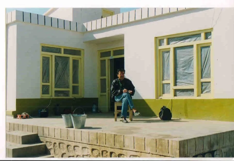 Χαμηλό λευκό σπίτι με τζάμια καλυμμένα με νάιλον· ένας άνδρας κάθεται στην τσιμεντένια βεράντα με τον εξοπλισμό δίπλα του.
