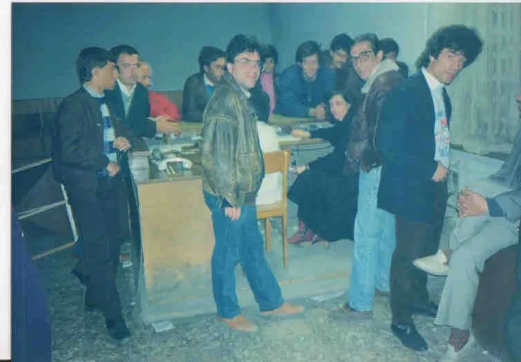 Γεμάτο δωμάτιο με άνδρες γύρω από γραφεία και χαρτιά, όρθιοι και καθισμένοι, σε κτίριο με γαλάζιους τοίχους.
