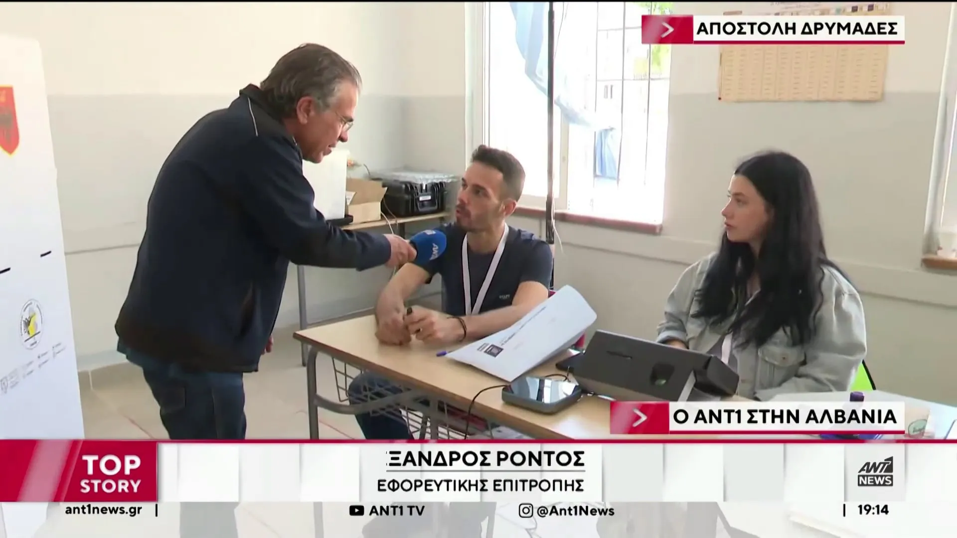 Ο Αργύρης Ντινόπουλος κρατά μικρόφωνο προς μέλος εφορευτικής επιτροπής καθισμένο σε σχολικό θρανίο εκλογικού τμήματος.