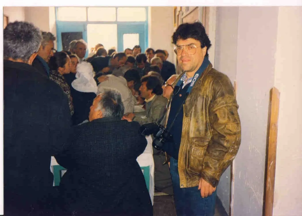 Πλήθος ψηφοφόρων γύρω από τραπέζια σε αίθουσα ψηφοφορίας· δεξιά ο Αργύρης Ντινόπουλος με δερμάτινο μπουφάν και φωτογραφική μηχανή.