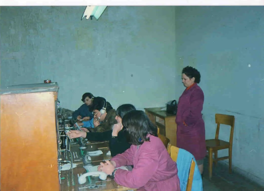 Αίθουσα με ανθρώπους σε γραφεία· δεξιά, γυναίκα στέκεται σε επιτοίχιο τηλέφωνο σε φθαρμένο εσωτερικό.