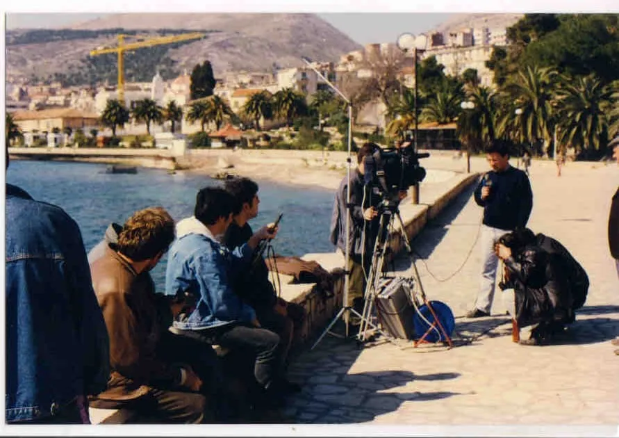 Δορυφορικό πιάτο και κάμερα σε τρίποδο στημένα σε παραλιακό μόλο, με το συνεργείο καθισμένο σε τοιχίο και τον κόλπο πίσω.