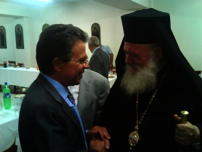 Ο Αργύρης Ντινόπουλος με κοστούμι σε θερμή χειραψία με ορθόδοξο ιεράρχη σε αίθουσα με εικόνες.