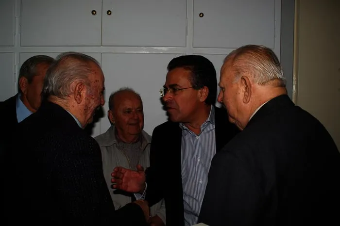 Ο Αργύρης Ντινόπουλος συνομιλεί με ομάδα ανδρών σε εσωτερικό χώρο.