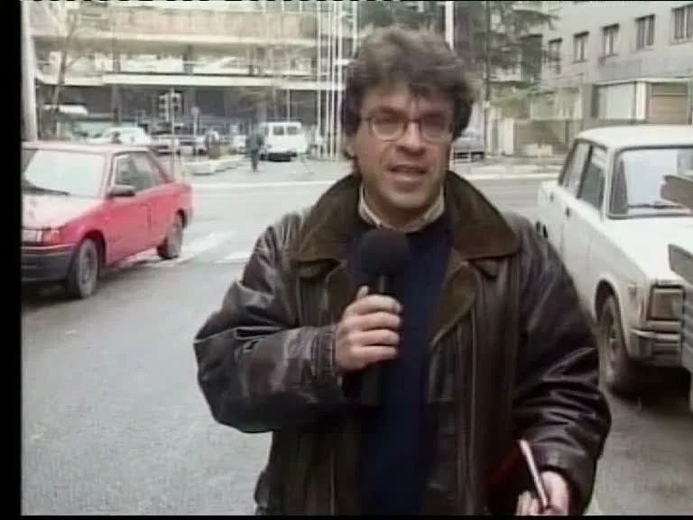 Ο Αργύρης Ντινόπουλος μιλά στην κάμερα με μικρόφωνο σε δρόμο του Βελιγραδίου, με σταθμευμένα αυτοκίνητα και πολυκατοικίες πίσω του, 1999.