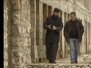 Ορθόδοξος μοναχός με μαύρα ράσα περπατά μαζί με έναν άνδρα κατά μήκος πέτρινου κτιρίου μονής, Κόσοβο, 2006.