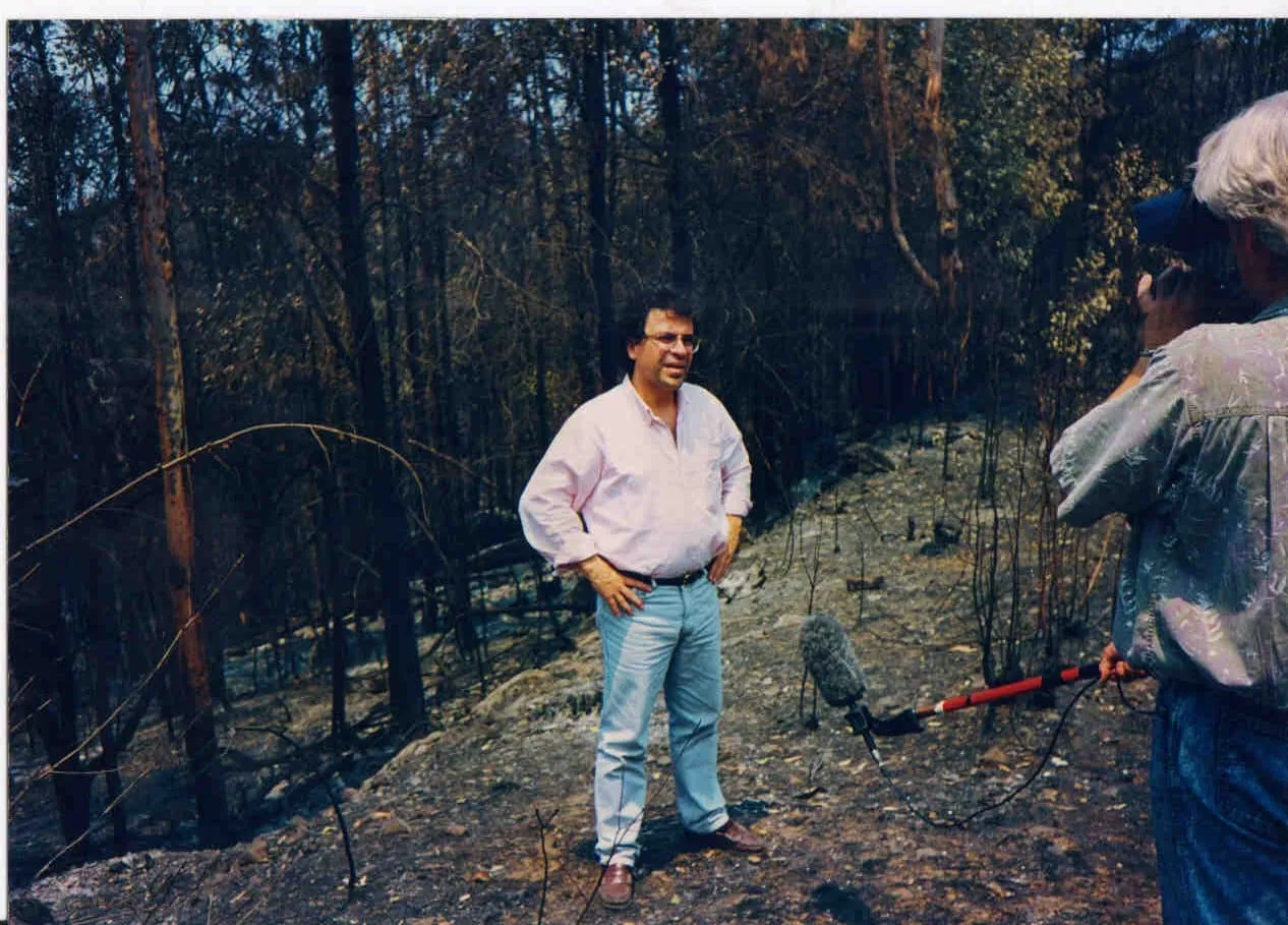 Argyris Dinopoulos standing in burnt forest, in a pink shirt with the sleeves rolled up, glasses and pale jeans, hands on his hips and his gaze off to the right; around him blackened trunks and branches, burnt ground, and a little green foliage higher up; at the right, close to the lens and cropped by the frame, someone with light hair seen from behind, in a pale patterned shirt and jeans, holding in one hand a red pole carrying a microphone in a fluffy cover, angled down and to the left, the other hand raised in front of them; beside their head a dark blue surface that cannot be made out.