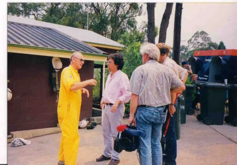 Outside a low building with dark red-brown walls and a corrugated roof, a man with light hair, dark glasses and a short-sleeved yellow boiler suit with a round badge on the arm is speaking and gesturing with both hands; facing him, Argyris Dinopoulos in a pink shirt and pale trousers; between them, seen from behind, two people, one of them wearing headphones, and low in the frame a black bag with a red part on it; a pale hat hangs under the eaves; in the background wheeled bins, a dark blue body with a red top, and trees.