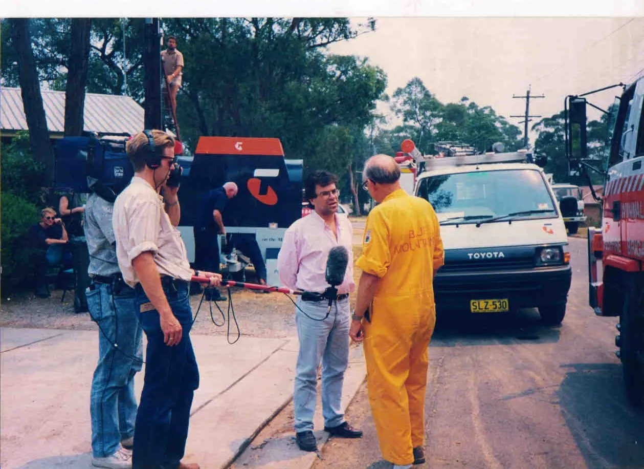 In a street with vehicles parked along it, Argyris Dinopoulos, in a pink shirt and glasses, is speaking to a man in a yellow boiler suit seen from behind, red lettering in two lines across his back, of which only «MOUNT…» can be read; raised between them, a microphone in a fluffy cover on a red pole, held from the left by someone wearing headphones; further back a television camera on a tripod and a person sitting in the shade; to the right a white van badged «TOYOTA» with the number plate «SLZ-530», and a red vehicle lettered «FIRE BRIGADE»; to the left a dark blue body with an orange top and a large orange emblem, and high above, a man on a ladder beside a pole, and at the blue body a man bent over it.
