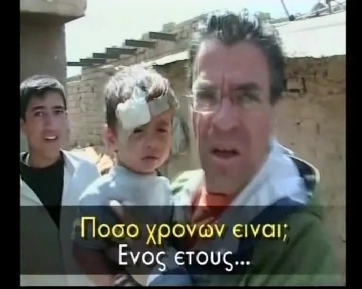 Ο Αργύρης Ντινόπουλος κρατά στην αγκαλιά του μικρό παιδί με επίδεσμο στο μέτωπο· δίπλα τους ένας έφηβος. Βαγδάτη, 2003. Υπότιτλος στο καρέ: «Πόσο χρονών είναι;» — «Ενός έτους...».