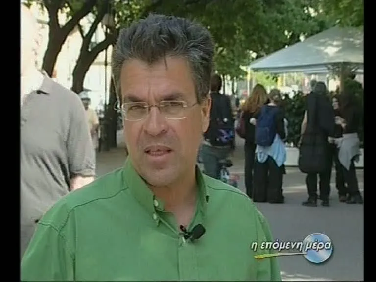 Frame from a television broadcast — Argyris Dinopoulos speaking to camera in an open, tree-shaded space, in a green shirt, thin-framed glasses and a clip-on microphone; at the left edge, cropped by the frame and out of focus, the back of someone passing; behind him passers-by, a blue backpack among them, and a white canopy to the right; lower right, the broadcast graphic «η επόμενη μέρα» — «the next day» — with a curving line beneath it running into a pale-blue disc.