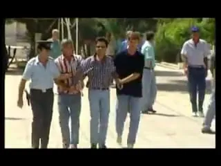 Καρέ από τηλεοπτικό υλικό, χαμηλής ανάλυσης. Ομάδα ανδρών προχωρά προς τον φακό σε ασφαλτοστρωμένο δρόμο. Στο κέντρο ένας άνδρας με ριγέ πουκάμισο βαδίζει ανάμεσα σε άλλους· αρκετοί φορούν ανοιχτογάλαζα κοντομάνικα πουκάμισα και δύο ένστολες φιγούρες στέκονται αριστερά και δεξιά πίσω. Δέντρα και χαμηλή βλάστηση στο βάθος.