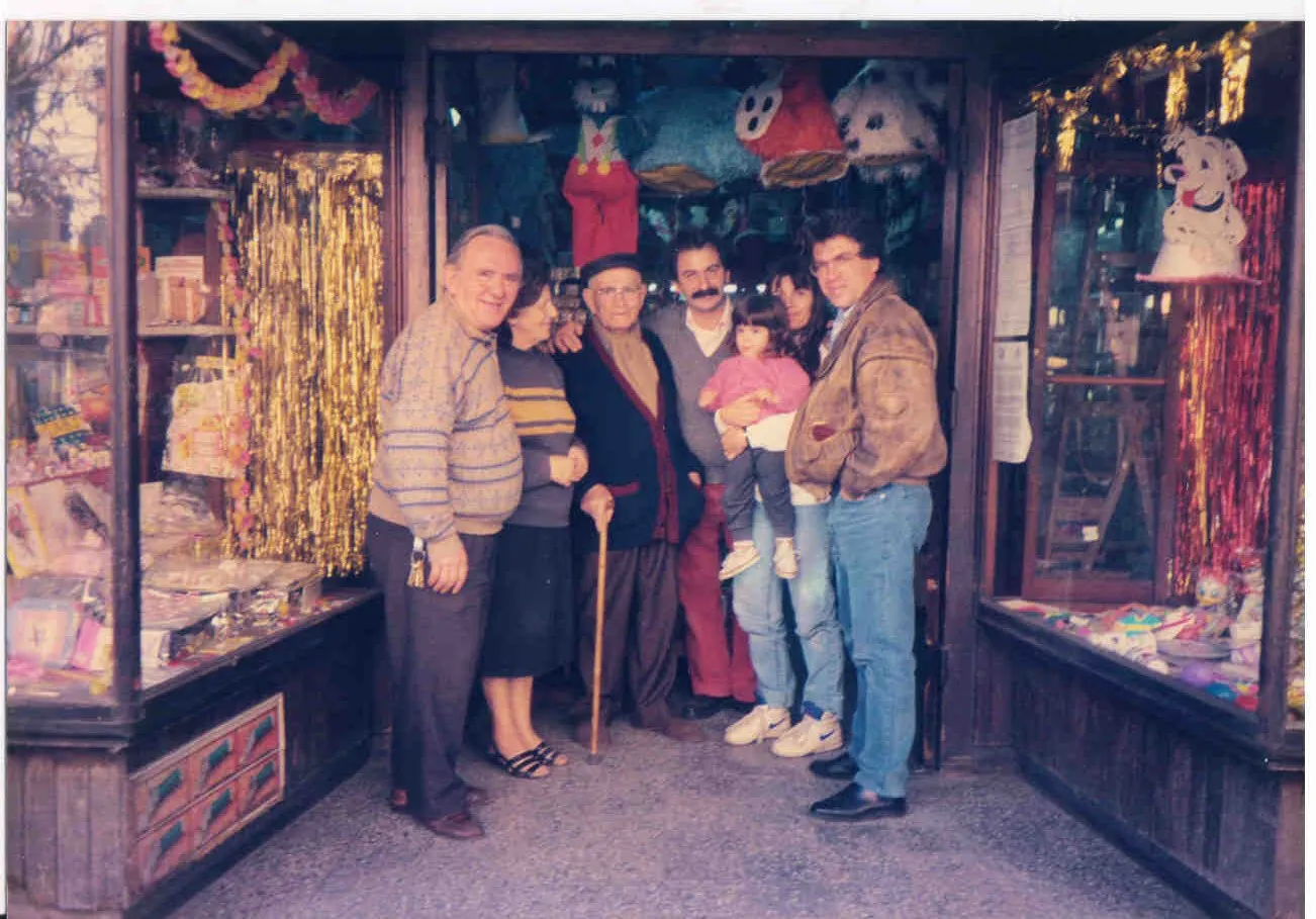 A group posing in the doorway of a sweet shop; on either side two large glass display cases full of confectionery and party goods, with gold and pink tinsel hanging and paper figures at the ceiling; at the centre an elderly man in a flat cap, maroon knitted cardigan and walking stick, beside him an older couple, a moustachioed man, a woman holding a small child in her arms, and at the right Argyris Dinopoulos in a leather jacket and jeans; upper right a cardboard Dalmatian figure.