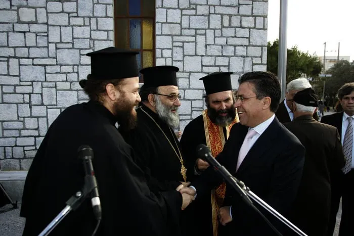 Ο Αργύρης Ντινόπουλος σε χειραψία με ορθόδοξο κλήρο έξω από πέτρινη εκκλησία, με μικρόφωνα μπροστά.
