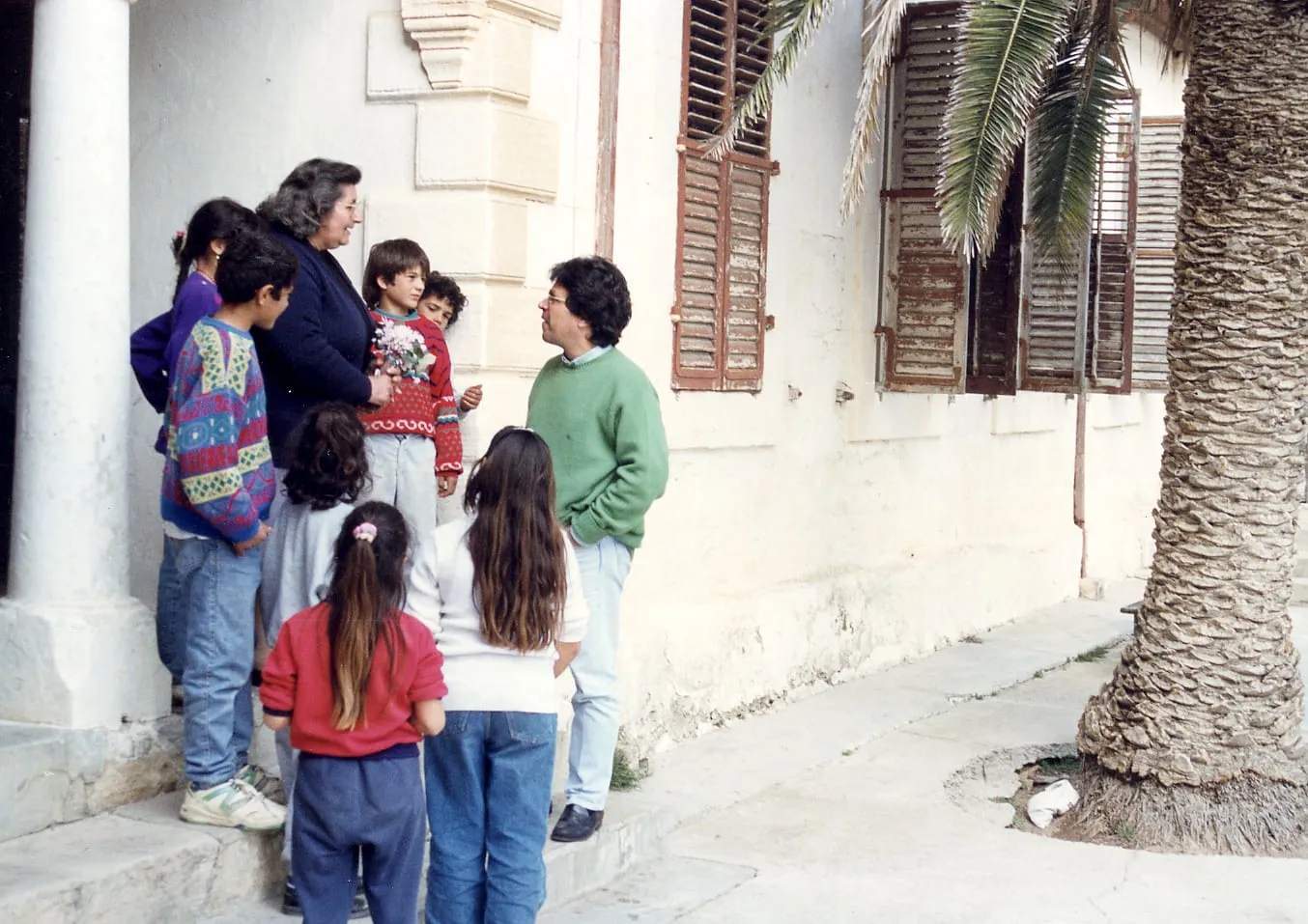 Ο Αργύρης Ντινόπουλος συνομιλεί με μια γυναίκα και επτά παιδιά έξω από ασβεστωμένο κτίριο με καφέ παντζούρια και φοίνικα δίπλα· η γυναίκα κρατά ανθοδέσμη.