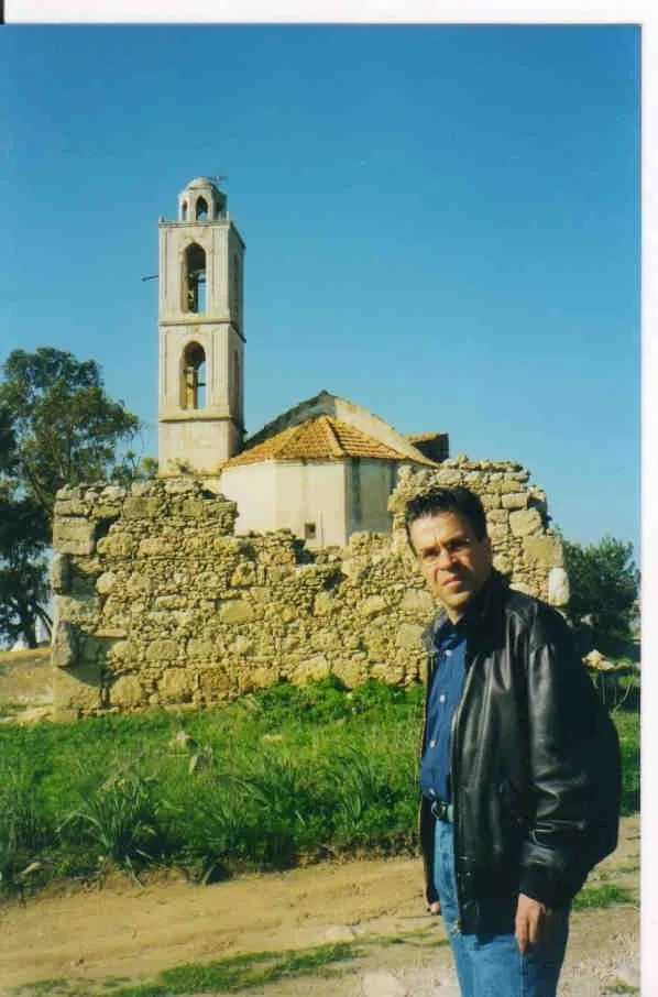 Ο Αργύρης Ντινόπουλος με δερμάτινο μπουφάν πάνω από τζιν πουκάμισο, μπροστά σε εκκλησία που στέκεται ακόμη πίσω από γκρεμισμένο πέτρινο περίβολο· το διώροφο καμπαναριό και η κεραμοσκεπή της είναι ακέραια.