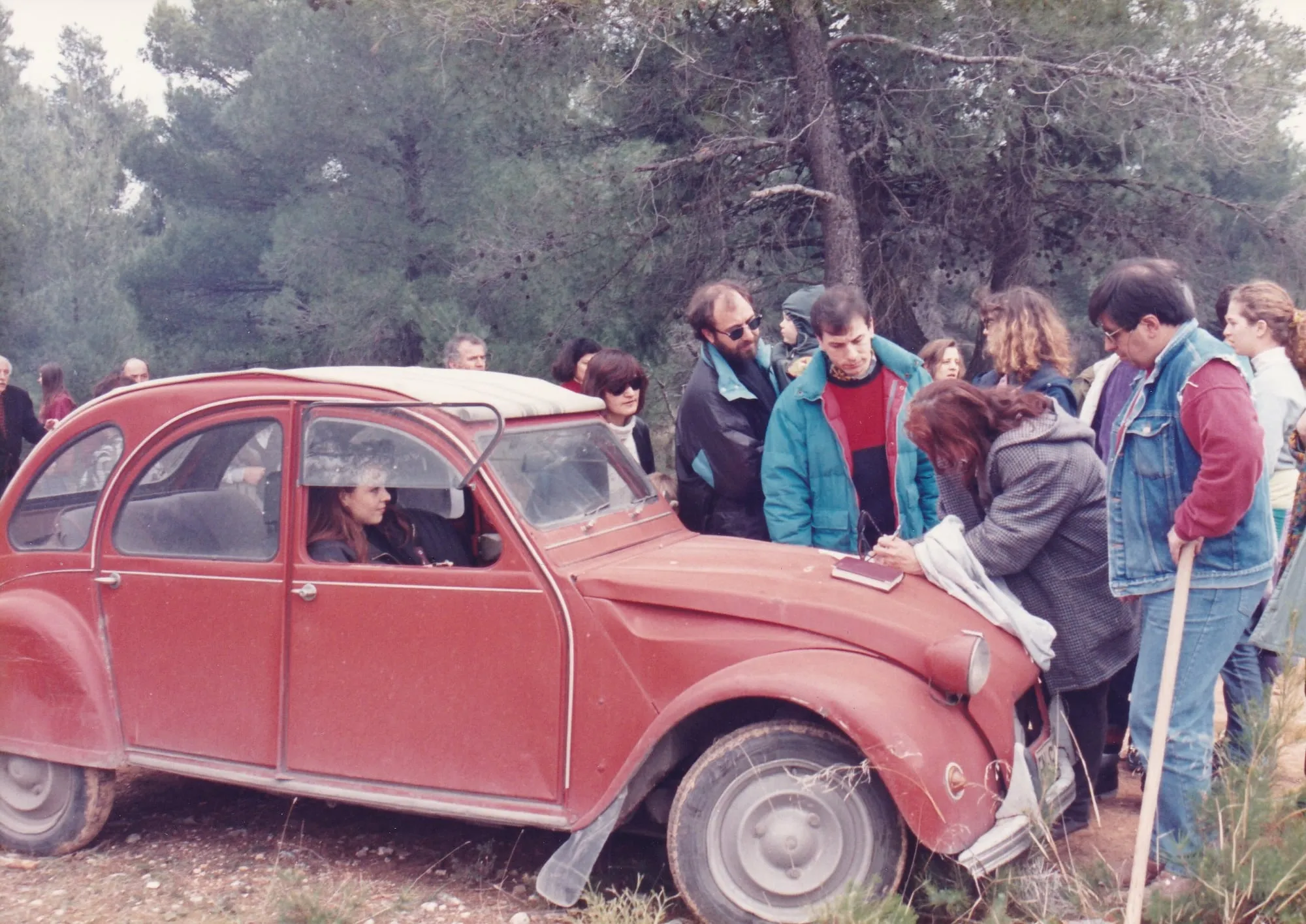 Υπαίθρια εκδήλωση δενδροφύτευσης σε πευκόφυτη πλαγιά με φρεσκοσκαμμένο χώμα. Στο κέντρο το κόκκινο Citroën 2CV του Αργύρη Ντινόπουλου με ανοιχτό καπό-γραφείο, όπου κάποιοι υπογράφουν σε χαρτιά· γύρω του άνθρωποι με χειμωνιάτικα μπουφάν, ανάμεσά τους ένα μικρό παιδί με πράσινο μπουφάν με κουκούλα, στην αγκαλιά ενήλικα. Στο βάθος άσπρα βαν και δέντρα.