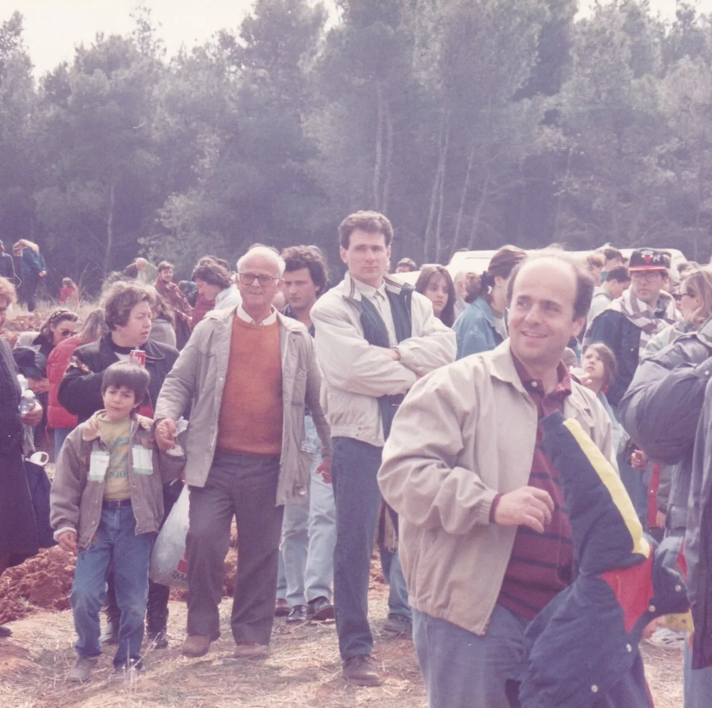 Πλήθος σε υπαίθρια εκδήλωση δενδροφύτευσης, με σκαμμένο χώμα μπροστά και πευκόδασος πίσω. Δεκάδες άνθρωποι με χειμωνιάτικα μπουφάν στέκονται και περπατούν προς τον φακό· ανάμεσά τους ένας ηλικιωμένος άνδρας με πορτοκαλί πουλόβερ και ανοιχτόχρωμη καμπαρντίνα, και δύο παιδιά — ένα αγόρι μπροστά αριστερά που κρατά το χέρι ενήλικα και φορά καρτελάκι εκδήλωσης, και ένα μικρότερο παιδί δεξιά του κέντρου ανάμεσα σε ενήλικες. Δεξιά διακρίνονται λευκά οχήματα.
