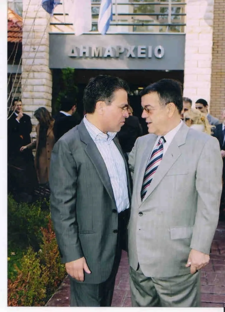 Δύο άνδρες με κοστούμι στέκονται κοντά ο ένας στον άλλο και μιλούν, σε πλακόστρωτο προαύλιο μπροστά από κτίριο με την επιγραφή «ΔΗΜΑΡΧΕΙΟ» και σημαίες από πάνω. Αριστερά ο Αργύρης Ντινόπουλος με γκρι κοστούμι και ριγέ πουκάμισο χωρίς γραβάτα, στραμμένος προς τα δεξιά· απέναντί του ένας μεγαλύτερος άνδρας με ανοιχτόχρωμο κοστούμι και σκούρα ριγέ γραβάτα. Πίσω τους αρκετοί άνθρωποι με επίσημο ντύσιμο και θάμνοι.