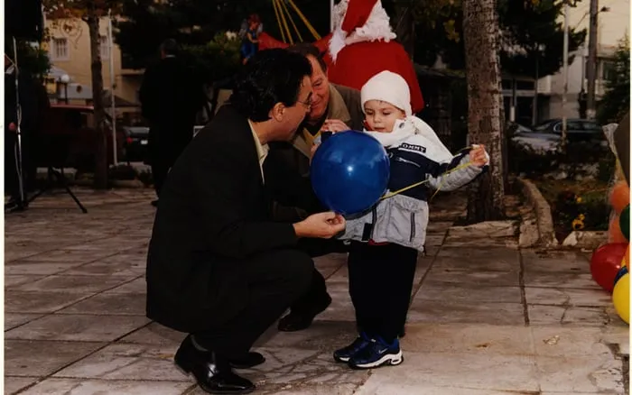 Ο Αργύρης Ντινόπουλος σκύβει να δώσει ένα μπαλόνι σε ένα παιδί σε γιορτινή πλατεία.
