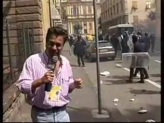 Καρέ από βίντεο — ο Αργύρης Ντινόπουλος μιλά στην κάμερα σε στενό δρόμο, με ριγέ ροζ-και-λευκό πουκάμισο και σηκωμένα μανίκια, σκούρο μικρόφωνο στο ένα χέρι και το άλλο υψωμένο, με μια κίτρινη κάρτα στο στήθος του· αριστερά του πέτρινο κτίριο με μεταλλικό κιγκλίδωμα, στη μέση του κάδρου ένας κατακόρυφος στύλος, και πιο πίσω στο οδόστρωμα ένα σταθμευμένο αυτοκίνητο, ένα ανοιχτόχρωμο όχημα, σκόρπια χαρτιά και μορφές με σκούρα ρούχα και κράνη — δύο από αυτές πίσω από μεγάλες διαφανείς ορθογώνιες ασπίδες.