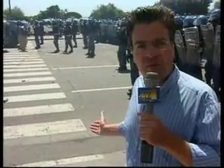 Καρέ από βίντεο — ο Αργύρης Ντινόπουλος μιλά στην κάμερα, δεξιά και πολύ κοντά στον φακό, με γαλάζιο ριγέ πουκάμισο, κρατώντας μικρόφωνο του οποίου το σκούρο μπλε πλαίσιο φέρει ένα χρυσαφί σήμα, και με το άλλο χέρι υψωμένο και τον αντίχειρα ανοιχτό· πίσω του, σε όλο το πλάτος του κάδρου, πυκνή παράταξη ανδρών με σκούρες στολές, ανοιχτόχρωμα κράνη και διαφανείς ασπίδες, σε φαρδύ δρόμο· μπροστά αριστερά μια διάβαση πεζών με λευκές λωρίδες, και πιο πίσω δέντρα και ανοιχτός ουρανός.