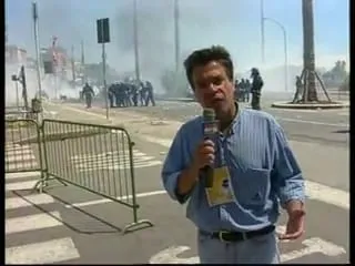 Καρέ από βίντεο — ο Αργύρης Ντινόπουλος μιλά στην κάμερα σε δρόμο με λευκές διαγραμμίσεις, με γαλάζιο ριγέ πουκάμισο, σκούρο μικρόφωνο στο χέρι και μια κίτρινη κάρτα κρεμασμένη από κορδόνι στο στήθος του· μπροστά αριστερά πράσινα μεταλλικά κιγκλιδώματα, άλλα στημένα και άλλα γερμένα στο οδόστρωμα· πιο πίσω καπνός απλωμένος χαμηλά, μια ομάδα μορφών με σκούρα ρούχα και κράνη μαζεμένων μεταξύ τους, μια μεμονωμένη μορφή αριστερά τους, ένα φανάρι σε στύλο, ένας ψηλός φανοστάτης και κτίρια που χάνονται στη θολούρα.