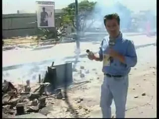 Καρέ από βίντεο — ο Αργύρης Ντινόπουλος στέκεται σε ανοιχτόχρωμο πλακόστρωτο με γαλάζιο πουκάμισο και μια κίτρινη κάρτα κρεμασμένη από κορδόνι στο στήθος του, κρατώντας υψωμένο ένα μικρό σκούρο αντικείμενο· μπροστά αριστερά απλώνονται καμένα και σπασμένα υπολείμματα, με ένα σκούρο όρθιο φύλλο ανάμεσά τους και καπνό να σέρνεται χαμηλά πάνω από το έδαφος· πίσω, ένας μακρύς γκριζοπράσινος φράχτης, και πάνω του μια μεγάλη ανοιχτόχρωμη αφίσα με σκούρα εικόνα μιας όρθιας μορφής· δεξιά της δέντρα και ένα ανοιχτόχρωμο αντικείμενο που δεν διακρίνεται.