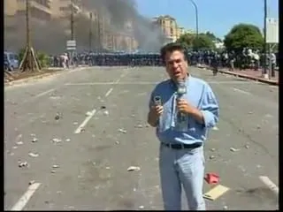 Καρέ από βίντεο — ο Αργύρης Ντινόπουλος στέκεται στο κέντρο ενός πλατιού οδοστρώματος με λευκές διαγραμμίσεις, με γαλάζιο πουκάμισο, ανοιχτόχρωμο τζιν και σκούρα ζώνη, κρατώντας μικρόφωνο με το καλώδιό του να κρέμεται και ένα μικρό σκούρο αντικείμενο στο άλλο χέρι· γύρω από τα πόδια του σκόρπιες ανοιχτόχρωμες πέτρες και μπάζα, ένα κόκκινο αντικείμενο και μια κίτρινη σανίδα· πίσω του μια μακριά σειρά μορφών με σκούρα ρούχα σε όλο το πλάτος του κάδρου, και πιο πίσω σκούρος καπνός που ανεβαίνει μπροστά από ένα μεγάλο πέτρινο κτίριο, δέντρα, ένας φανοστάτης και γαλάζιος ουρανός· αριστερά ένα νεαρό δέντρο σε τρίγωνο ξύλινο στήριγμα και ένα ανοιχτόχρωμο όχημα πίσω του.