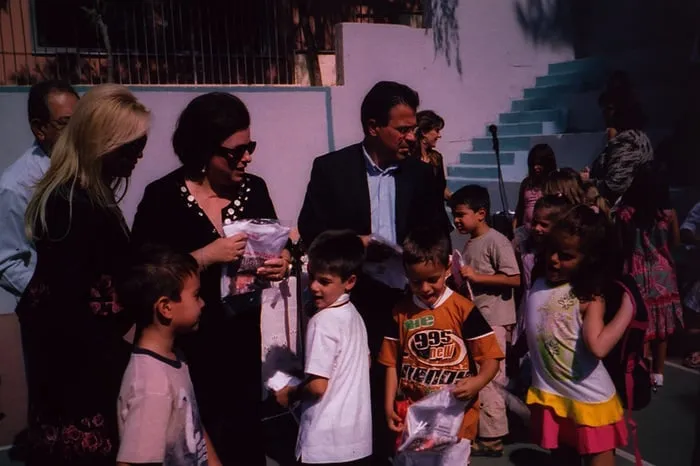 Δύο γυναίκες και ένας άνδρας μοιράζουν σακούλες με δώρα σε παιδιά σε αυλή με μπλε πλακόστρωτα σκαλιά.