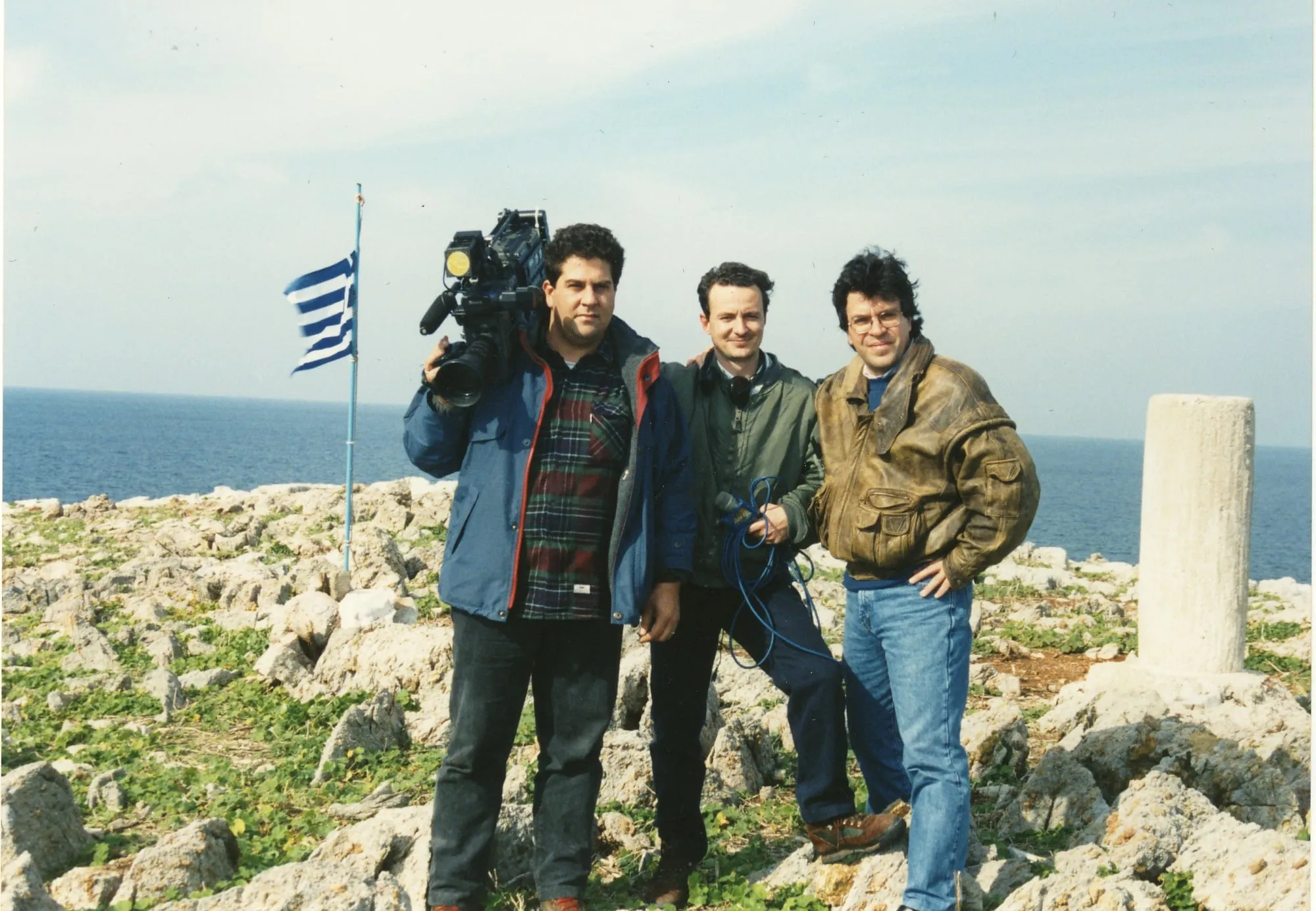 Τρεις άνδρες στη βραχώδη νησίδα: ένας με κάμερα στον ώμο, ένας με ακουστικά περασμένα στον λαιμό που κρατά μικρόφωνο και τυλιγμένο μπλε καλώδιο, και ο Αργύρης Ντινόπουλος· ελληνική σημαία πίσω και λευκό κτιστό ορόσημο δίπλα τους.