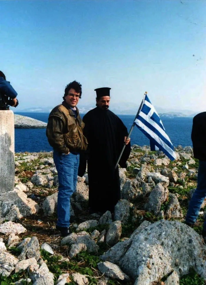 Ο ανταποκριτής Αργύρης Ντινόπουλος με ορθόδοξο ιερέα που κρατά ελληνική σημαία σε βραχονησίδα, με τη θάλασσα πίσω, Ίμια, 1996.