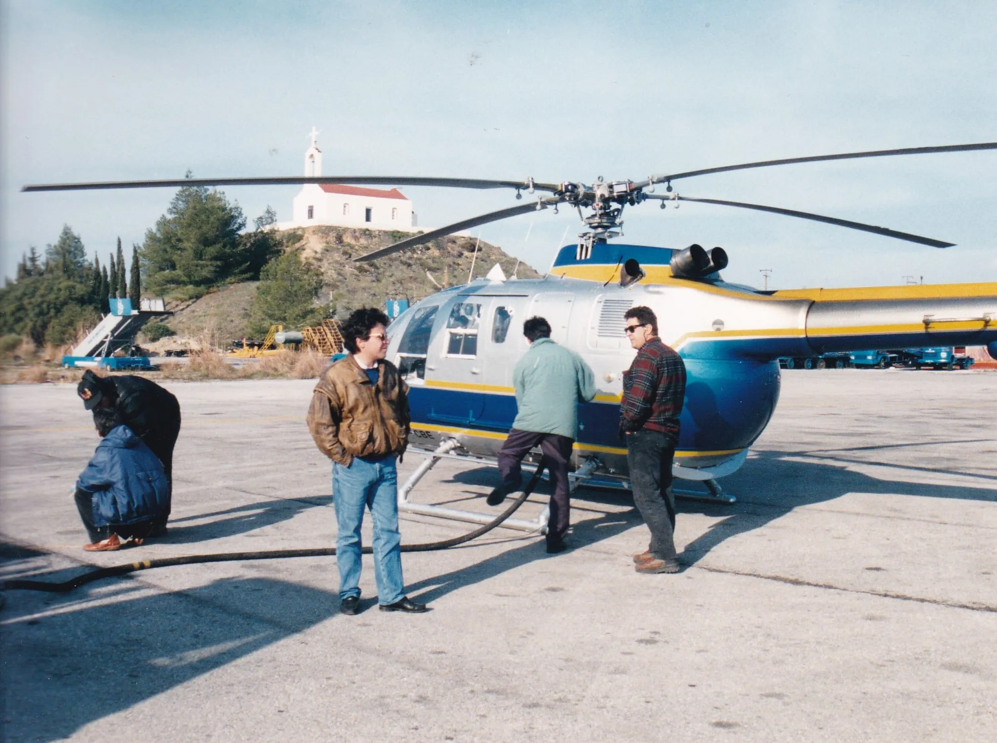 Το ελικόπτερο ανεφοδιάζεται με καύσιμα σε δάπεδο αεροδρομίου, με λευκό εκκλησάκι σε λόφο πίσω και άνδρες γύρω από το ελικόπτερο, ο ένας σκυμμένος.