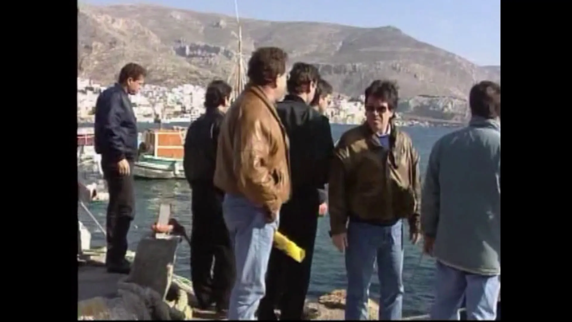 Ομάδα ανδρών στην προκυμαία, με τα σπίτια της Καλύμνου στην πλαγιά και σκάφη δεμένα πίσω τους.