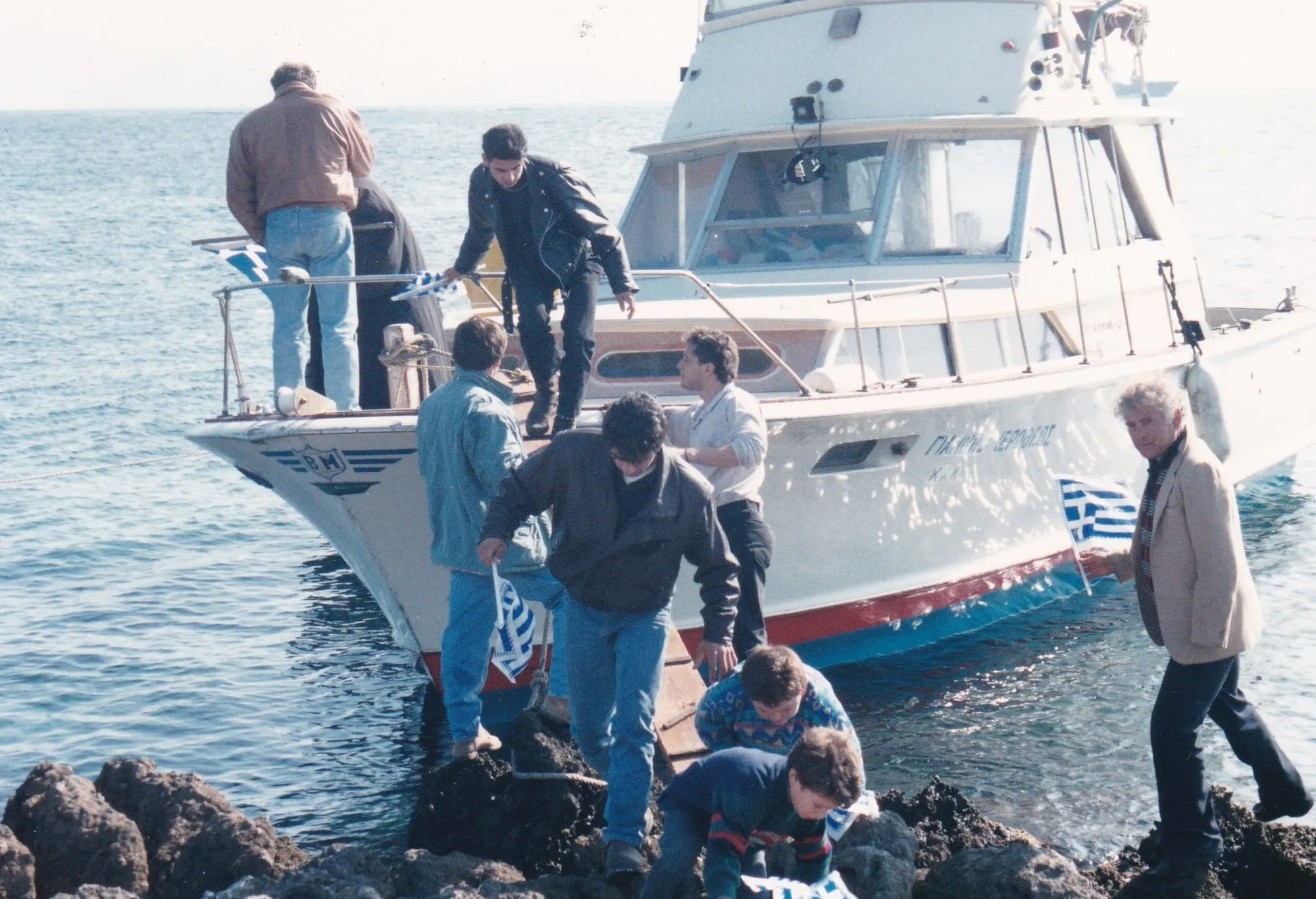 Άνθρωποι αποβιβάζονται στα βράχια από το λευκό σκάφος, στο κύτος του οποίου διαβάζεται «ΓΙΑΝΝΗΣ ΙΕΡΟΘΕΟΣ». Στο πρώτο πλάνο δύο άνδρες σκυμμένοι πάνω στα βράχια, με μικρές ελληνικές σημαίες δίπλα τους· δεξιά ένας άνδρας κρατά ανοιχτή σημαία σε κοντάρι.