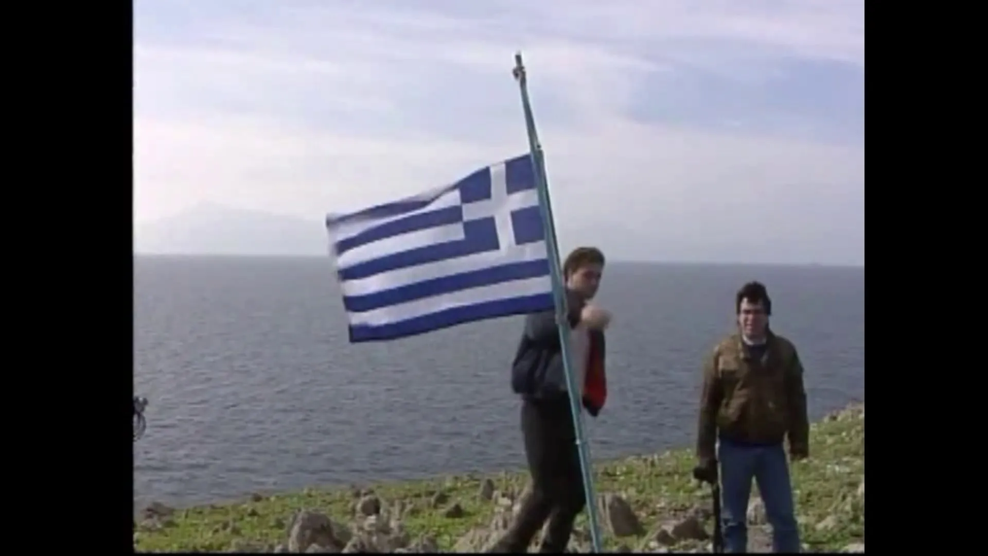 Ελληνική σημαία σε πρόχειρο ιστό στη βραχώδη ράχη της νησίδας, με δύο άνδρες δίπλα της και τη θάλασσα πίσω.