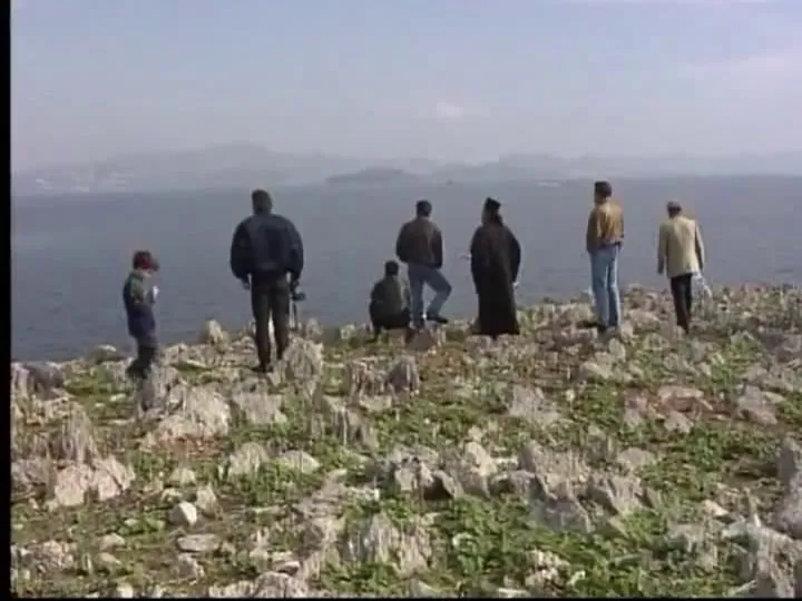 Επτά περίπου άνθρωποι σκορπισμένοι στη βραχώδη ράχη της νησίδας, ανάμεσά τους ιερέας με μαύρο ράσο και ένα παιδί.